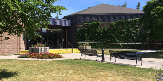 Entrance of a brick community building with yellow steps, a ramp with a railing, and ivy-covered walls. Benches and planters are nearby.