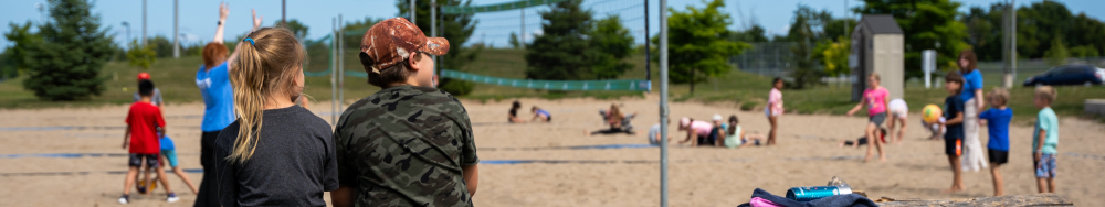 A group of people, including children and adults, at an outdoor sandy area that appears to be a beach or playground. Various activities are taking place, such as playing with balls and sitting on the sand. The background includes trees, a fence, and some structures.